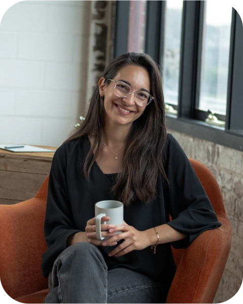 A photo of Ilaria, the owner of La Ria Creations. She has a bright, big smile on her face, while sitting on a chair and holding a cup of tea in her hand.